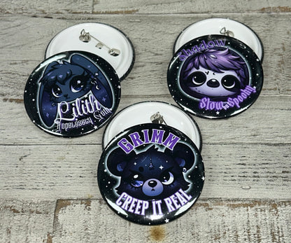 A set of 1.25-inch and 2.25-inch button pins featuring designs from the Grimm & Friends Goth Collection, including Grimm with "Creep it Real," a Goth Sloth with "Slow and Spooky," and Lilith the Goth Bunny with "Hopelessly Goth."