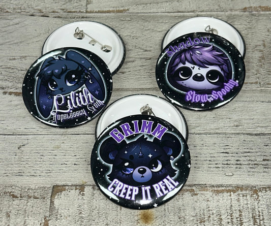 A set of 1.25-inch and 2.25-inch button pins featuring designs from the Grimm & Friends Goth Collection, including Grimm with "Creep it Real," a Goth Sloth with "Slow and Spooky," and Lilith the Goth Bunny with "Hopelessly Goth."